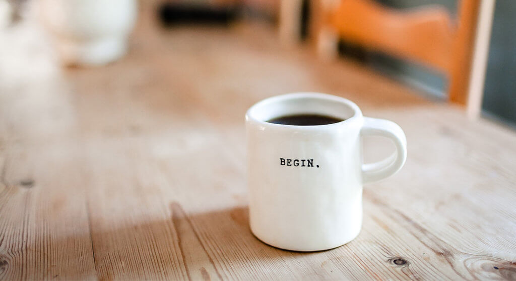 Coffee cup on a wooden table. The coffee cup has the word "begin" on it.