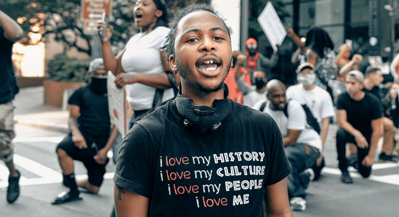 Black young man speaking out at a George Floyd Protest in Summer of 2020. He's wearing a tshirt that says "I love my history. I love my culture. I love my people. I love me." People of different racial and ethnic backgrounds are in the background behind him, some on one knee.