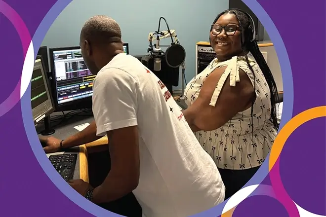 Smiling Black woman in a recording studio with a Black man adjusting details on the computer.